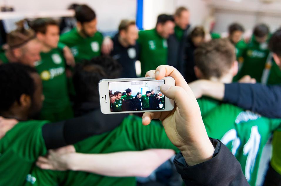 Hand hält ein Smartphone, das ein Team in grünen Trikots aufnimmt, während die Spieler einander umarmen.